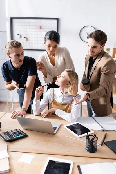 Femme blonde assise à table avec des employés de bureau sceptiques et multiculturels debout derrière, gesticulant et regardant un ordinateur portable sur le lieu de travail — Photo de stock