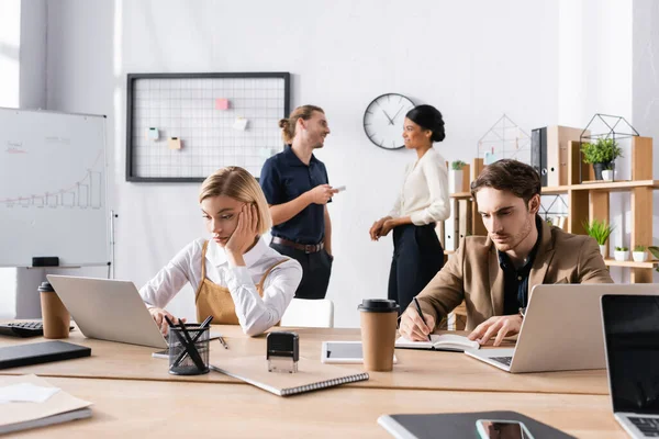 Los trabajadores de oficina aburridos utilizando el ordenador portátil y la escritura en el cuaderno mientras están sentados en la mesa, con colegas multiculturales hablando en segundo plano - foto de stock