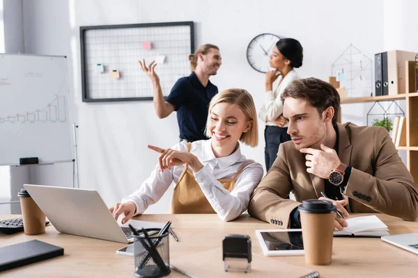 Hombre enfocado sentado en el lugar de trabajo cerca de la mujer señalando con el dedo, mientras que el uso de ordenador portátil con trabajadores de oficina multiculturales en segundo plano - foto de stock