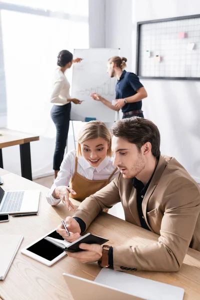 Excited woman looking at notebook in hand of man sitting at workplace in office, with blurred multicultural colleagues on background — Stock Photo