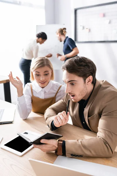 Excited office workers looking at notebook while sitting at table in office with blurred multicultural colleagues working on background — Stock Photo