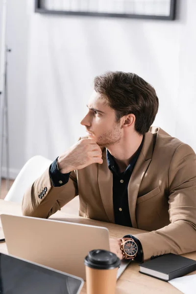 Thoughtful office worker looking away while sitting at workplace with laptop on blurred background — Stock Photo