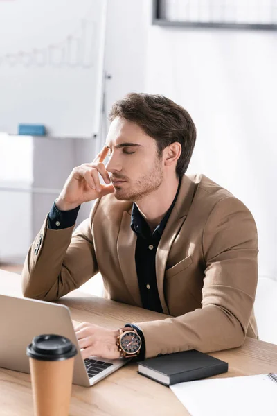 Empresário atencioso com olhos fechados segurando a mão perto da cabeça enquanto sentado na mesa com laptop no fundo embaçado — Fotografia de Stock