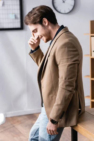 Vista lateral del hombre reflexivo con los ojos cerrados sonriendo mientras se apoya en el escritorio en la oficina con fondo borroso - foto de stock