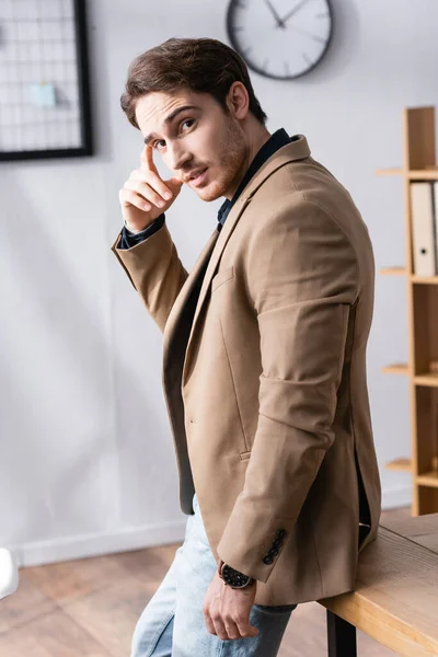 Thoughtful man hand near head looking at camera while leaning on desk in office on blurred background — Stock Photo