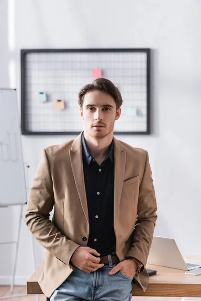 Front view of confident businessman with hand in pocket looking at camera while leaning on desk on blurred background — Stock Photo