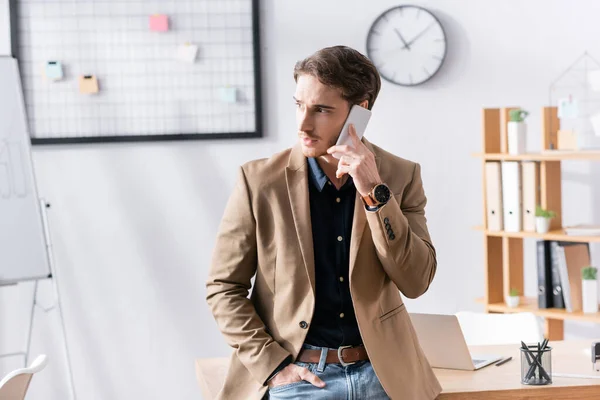 Homem confiante com a mão no bolso, olhando para o lado enquanto fala ao telefone, apoiando-se na mesa no escritório no fundo turvo — Fotografia de Stock