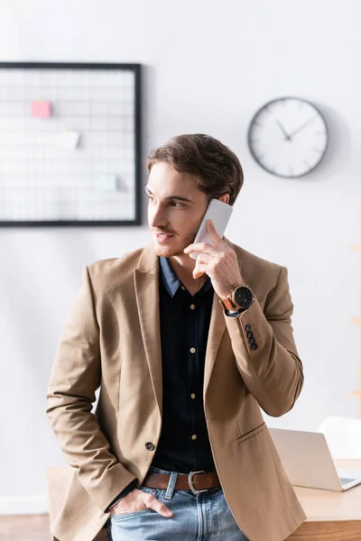 Hombre con la mano en el bolsillo, sonriendo mientras habla por teléfono, apoyado en la mesa en el lugar de trabajo sobre fondo borroso - foto de stock