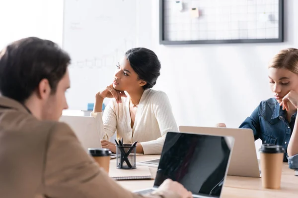 Gelangweilte Frauen mit Händen in Kopfnähe mit Laptops, während sie am Arbeitsplatz mit verschwommenem Mann im Vordergrund sitzen — Stockfoto