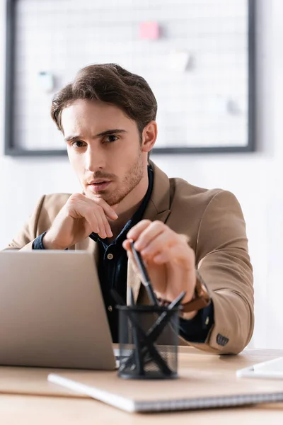 Retrato del trabajador de oficina confiado con pluma, mirando a la cámara mientras está sentado en la mesa con el ordenador portátil en primer plano borroso - foto de stock