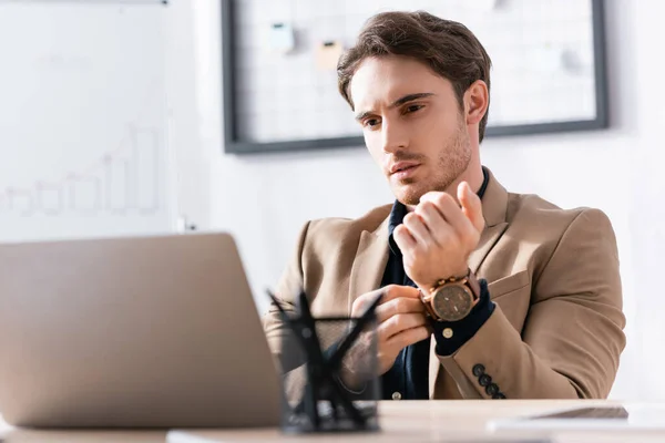 Концентрований бізнесмен випрямляє рукав куртки, дивлячись на ноутбук і сидячи на робочому столі на розмитому передньому плані — стокове фото