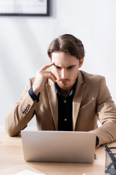 Thoughtful young adult man with hand near head looking at laptop while sitting at desk on blurred background — Stock Photo