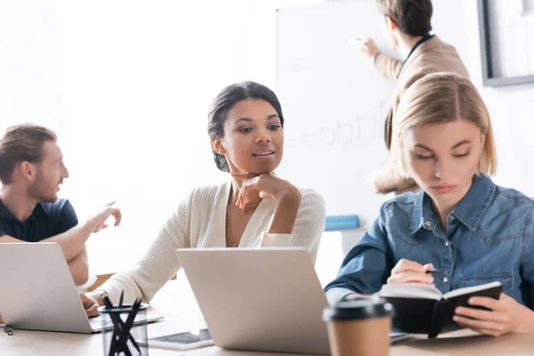 Positive african american woman looking at notebook in hands of colleague sitting near with blurred co-workers on background — Stock Photo