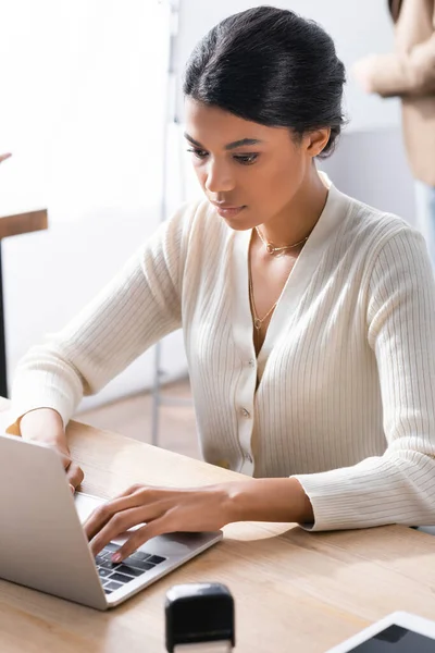 Konzentrierte Afroamerikanerin tippt auf Laptop, während sie am Schreibtisch auf verschwommenem Hintergrund sitzt — Stockfoto