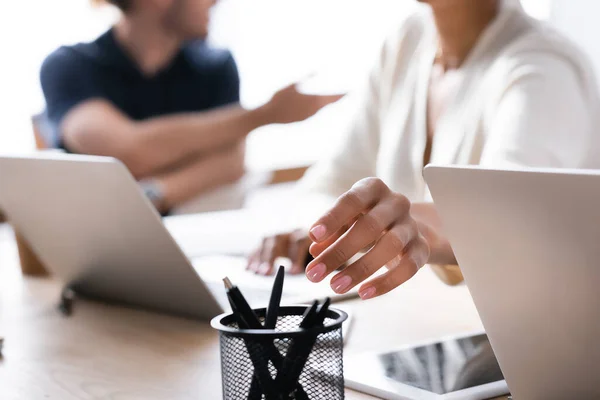 Cropped view of hand of african american woman taking pen from holder on desk with blurred man on background — Stock Photo