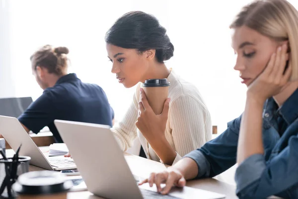 Focused african american woman with paper cup looking at laptop, while sitting at table with blurred tired woman on foreground — Stock Photo