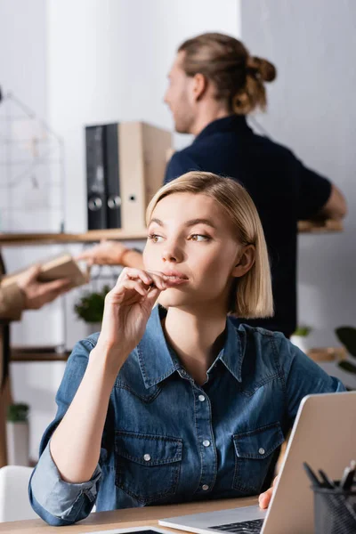 Thoughtful blonde woman looking away while sitting near laptop at workplace with blurred men on background — Stock Photo