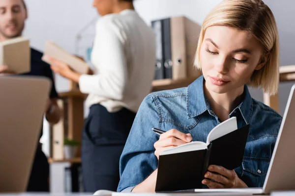 Blonde woman writing on notebook in office with blurred multicultural co-workers on background — Stock Photo