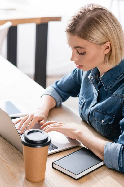 Overhead view of blonde woman typing on laptop while sitting near paper cup and notebook on table on blurred background — Stock Photo