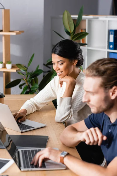 Smiling african american woman looking at laptop while sitting at workplace with blurred man on foreground — Stock Photo
