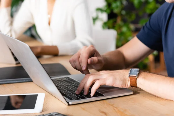 Ausgeschnittene Ansicht eines Mannes mit tippender Uhr auf dem Laptop, während er am Schreibtisch sitzt, mit verschwommener afrikanisch-amerikanischer Frau im Hintergrund — Stockfoto