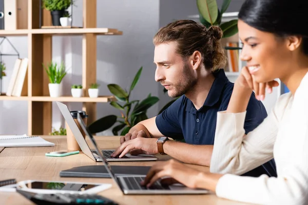 Focused young adult man looking at laptop while sitting at workplace with blurred african american woman on foreground — Stock Photo