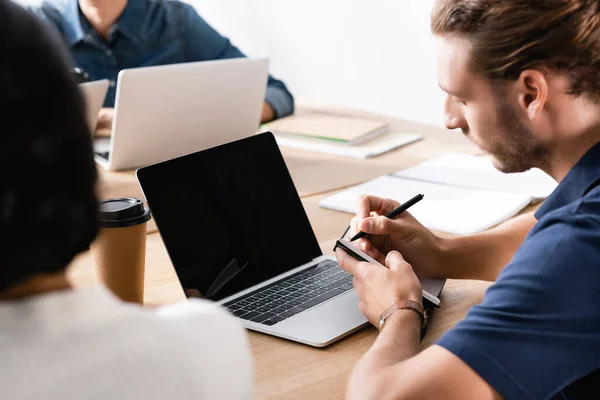 Trabajador de oficina escribiendo en un cuaderno mientras está sentado cerca de la computadora portátil con pantalla en blanco en el lugar de trabajo con la mujer afroamericana borrosa en primer plano - foto de stock