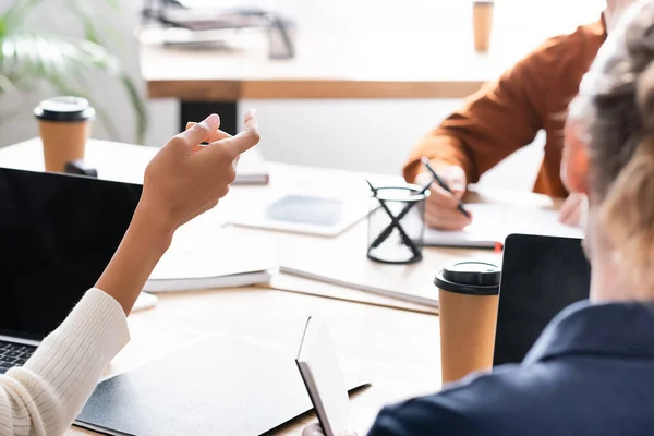 Vista ritagliata di donna afro-americana gesticolare mentre seduto sul posto di lavoro con sfocato co-lavoratore scrittura su sfondo — Foto stock