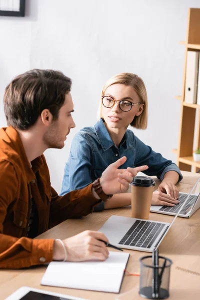 Mujer rubia en gafas mirando gestos de compañeros de trabajo, mientras está sentada en el escritorio con computadoras portátiles en la oficina sobre un fondo borroso - foto de stock