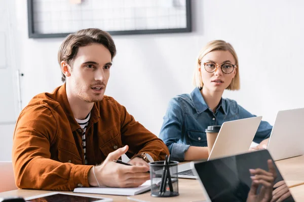 Donna bionda seduta sul posto di lavoro vicino all'uomo che parla e fa gesti in ufficio su sfondo sfocato — Foto stock