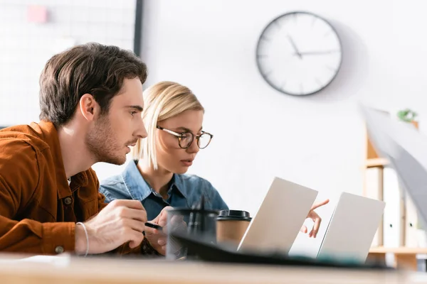 Hombre enfocado mirando a la computadora portátil, mientras está sentado cerca de la mujer señalando con el dedo en la oficina en primer plano borroso - foto de stock