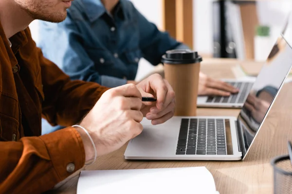 Ausgeschnittene Ansicht eines Mannes mit Stift, der auf den leeren Bildschirm seines Laptops blickt, während er am Arbeitsplatz sitzt, mit verschwommener Frau im Hintergrund — Stockfoto