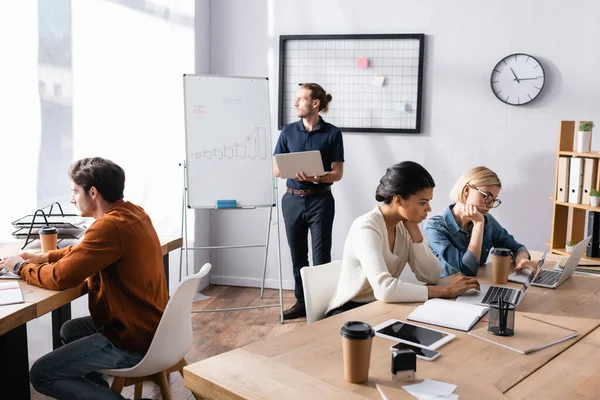 Young adult man with laptop looking away, while standing near multicultural co-workers sitting at workplaces in office — Stock Photo