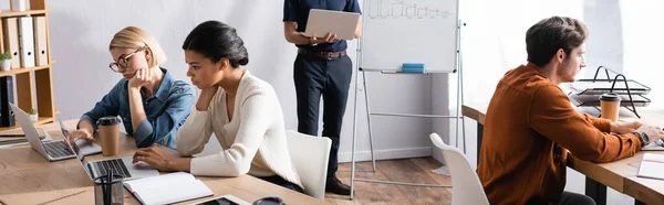Uomo in possesso di laptop e in piedi vicino a colleghi multiculturali seduti nei luoghi di lavoro in ufficio, banner — Foto stock
