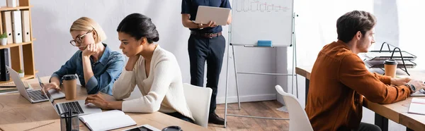 Man holding laptop and standing near multicultural colleagues sitting at workplaces in office, banner — Stock Photo