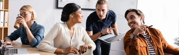 Mujer afroamericana sonriente mirando a sus compañeros de trabajo mientras está sentada en el escritorio cerca de un colega tomando café en la oficina, pancarta - foto de stock