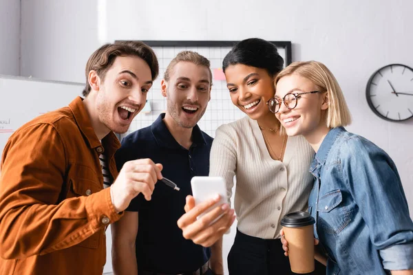 Aufgeregter Geschäftsmann zeigt mit Stift auf Smartphone in der Nähe multiethnischer Kollegen — Stockfoto