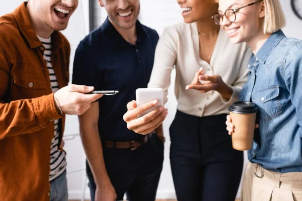 Cheerful businesspeople pointing with finger and pen and smartphone near multicultural colleagues on blurred background — Stock Photo