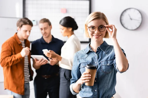 Sonriente gerente tocando anteojos y sosteniendo café para ir mientras colegas multiétnicos discutiendo sobre fondo borroso - foto de stock