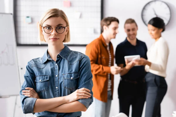 Joven, rubia empresaria de pie con los brazos cruzados mientras colegas multiétnicos hablando sobre fondo borroso - foto de stock