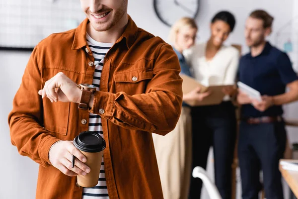 Sonriente hombre de negocios sosteniendo café para ir y mirando reloj de pulsera cerca de colegas hablando sobre fondo borroso — Stock Photo