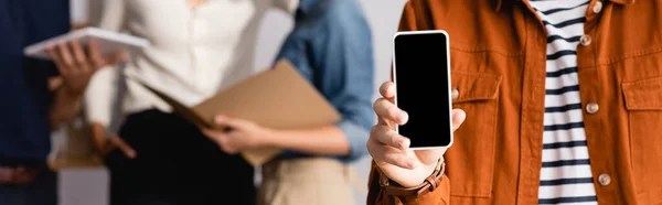 Vista parziale di uomo d'affari che mostra smartphone con schermo vuoto vicino a colleghi sfocati sullo sfondo, banner — Stock Photo
