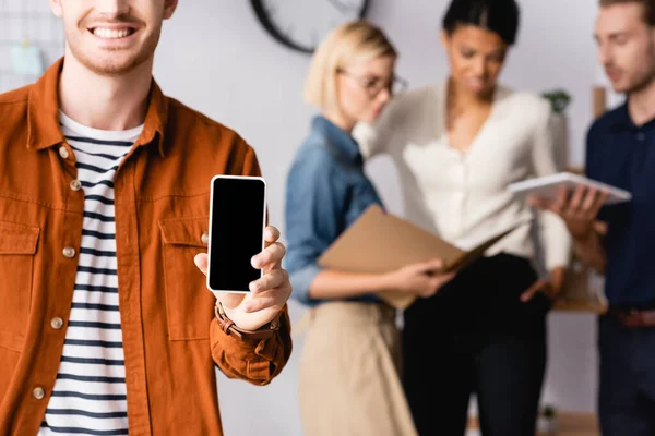 Gestor sonriente mostrando teléfono inteligente con pantalla en blanco cerca de empresarios multiétnicos que se comunican sobre un fondo borroso - foto de stock