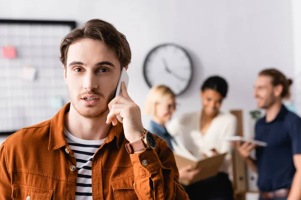 Jungunternehmer spricht auf Smartphone, während multikulturelle Kollegen über Projekt auf verschwommenem Hintergrund diskutieren — Stockfoto