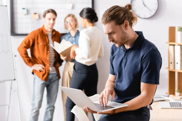 Sérieux gestionnaire tapant sur ordinateur portable près de gens d'affaires multiethniques discuter du projet sur fond flou — Photo de stock