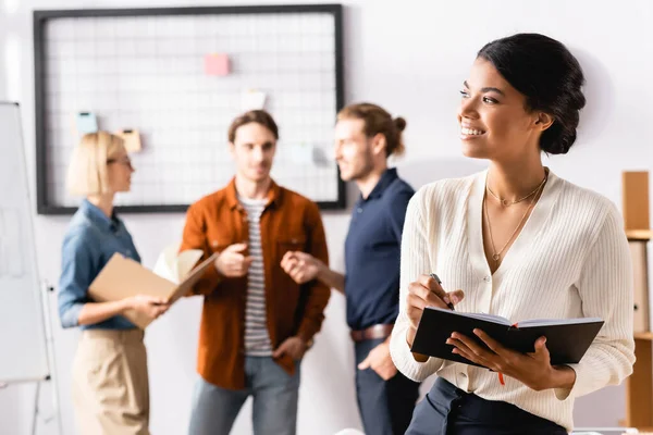 Feliz mujer de negocios afroamericana mirando hacia otro lado mientras colegas multiculturales se comunican sobre un fondo borroso - foto de stock