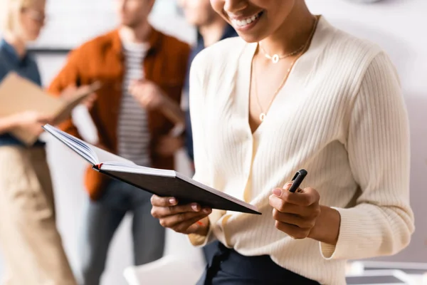 Cropped view of african american businesswoman holding notebook and pen near colleagues on blurred background — Stock Photo