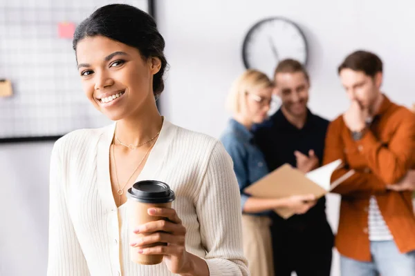 Happy african american businesswoman looking at camera while multicultural colleagues talking on blurred background — Stock Photo