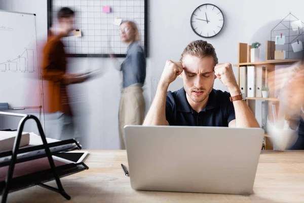 Homme d'affaires fatigué assis avec les mains sur la tête et les yeux fermés près d'un ordinateur portable dans un bureau ouvert, flou de mouvement — Photo de stock