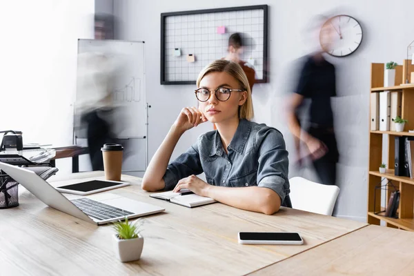 Femme d'affaires blonde en lunettes assis sur le lieu de travail avec des gadgets dans un bureau ouvert, flou de mouvement — Photo de stock